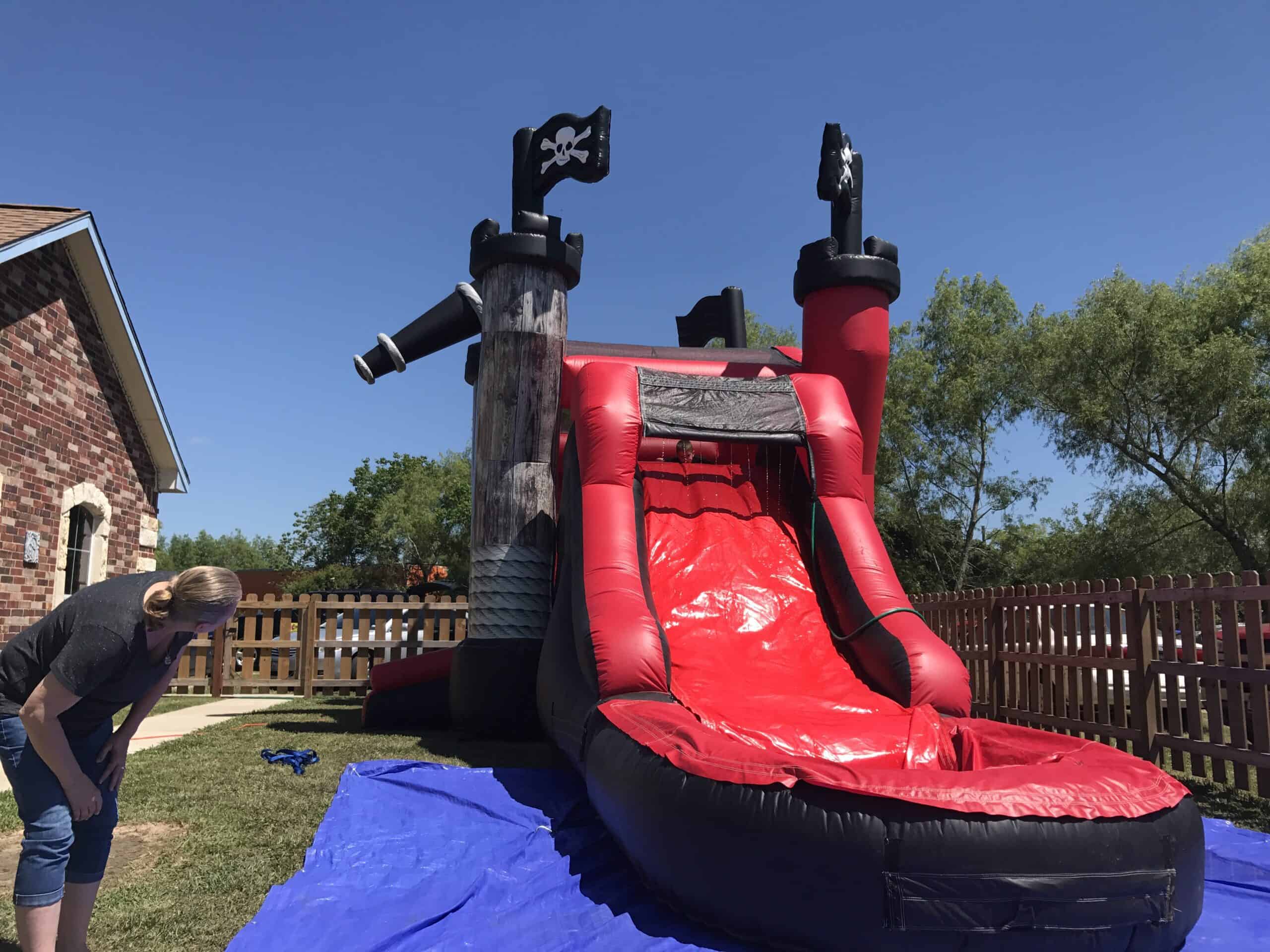 Red and black pirate ship inflatable water slide setup in a backyard with a blue ground tarp