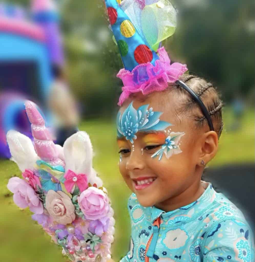 Children’s Floral Face Painting & Props Young girl smiling with floral face paint, a party hat, and holding a decorated unicorn wand