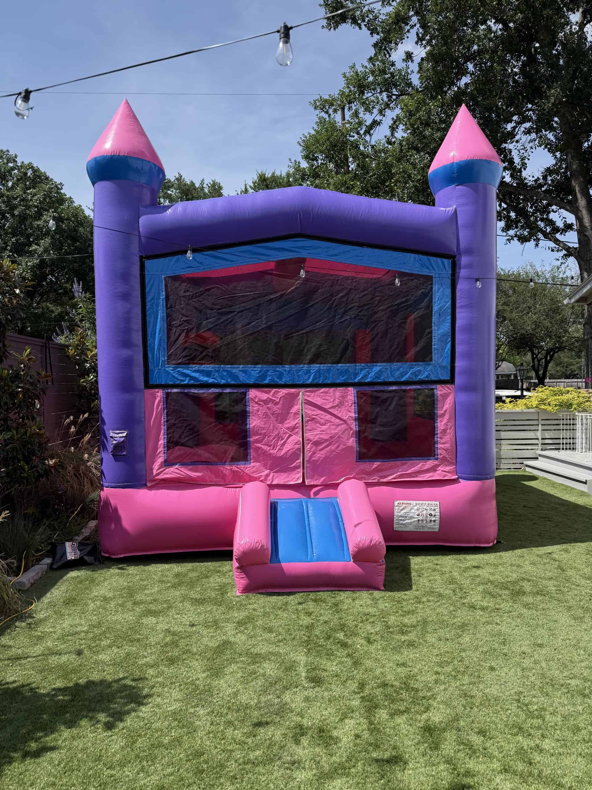 Pink and purple inflatable castle bounce house with a front entrance slide on green lawn.