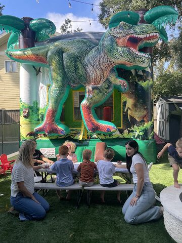 Colorful T-Rex bounce house with children playing and adults supervising outdoors.