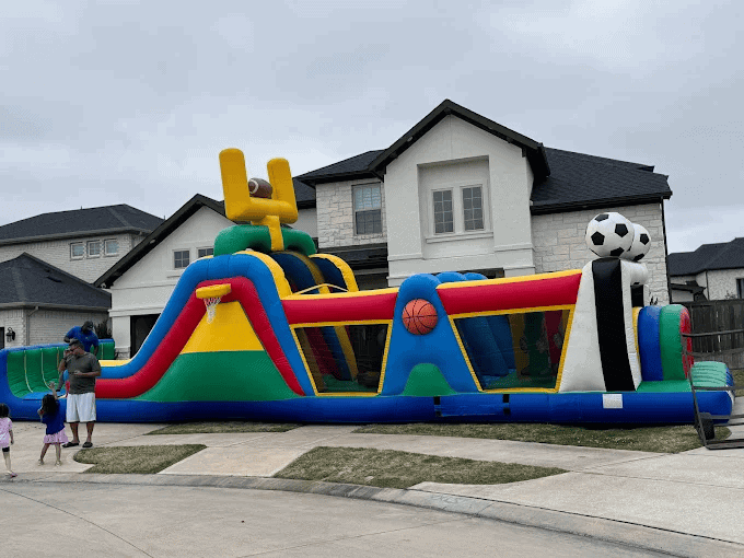 Colorful Sports Obstacle Course Bounce House Large inflatable sports obstacle course bounce house in front of a house.